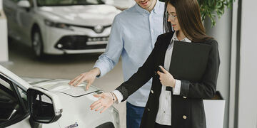 A man and a woman stand beside a white hybrid car in a showroom. The woman, holding a folder, gestures toward the car as the man touches its hood, both appearing engaged in conversation.