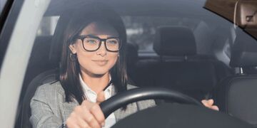 A woman wearing glasses and a blazer is sitting in the driver's seat of a car, holding the steering wheel and looking forward through the windscreen.
