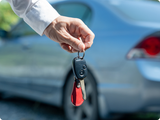 A person’s hand holding a car key fob and red keyring in front of a blurred blue car, suggesting car ownership or a car handover.